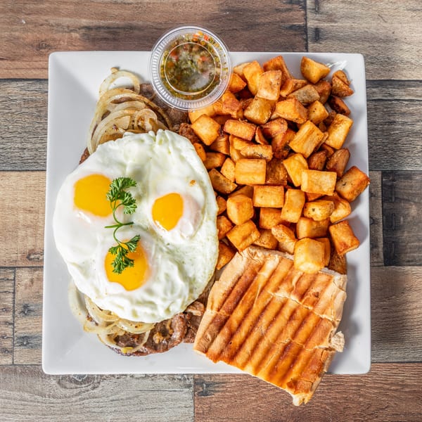 Cuban breakfast with steak, fried eggs, sautéed onions, home fries, and toasted bread on a white plate.