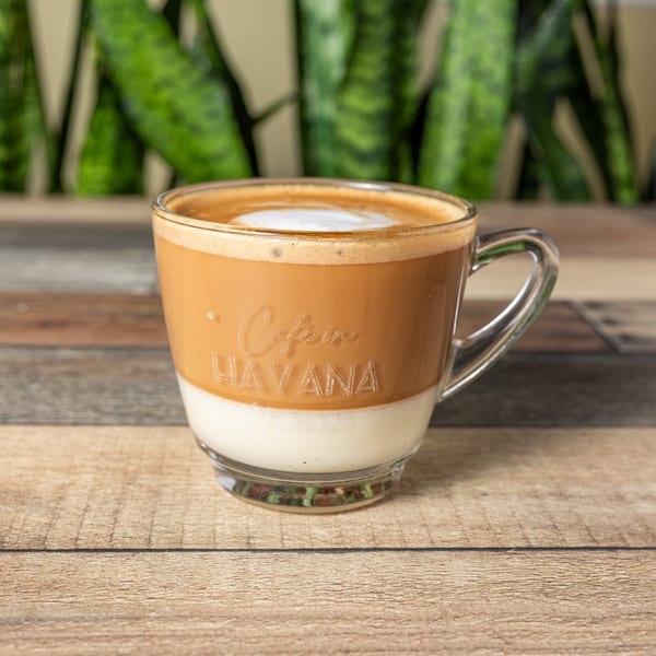 A clear glass mug of Café In Havana coffee with layered milk and foam on a wooden table.
