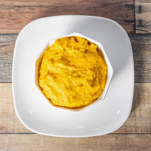 Overhead view of a white bowl filled with bright yellow mashed plantains on a white square plate.