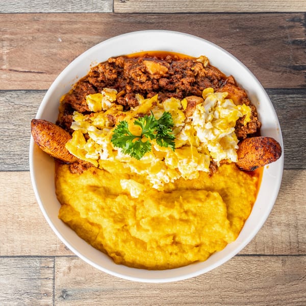 A Cuban breakfast bowl with picadillo, scrambled eggs, plantains, and yellow grits, garnished with parsley.
