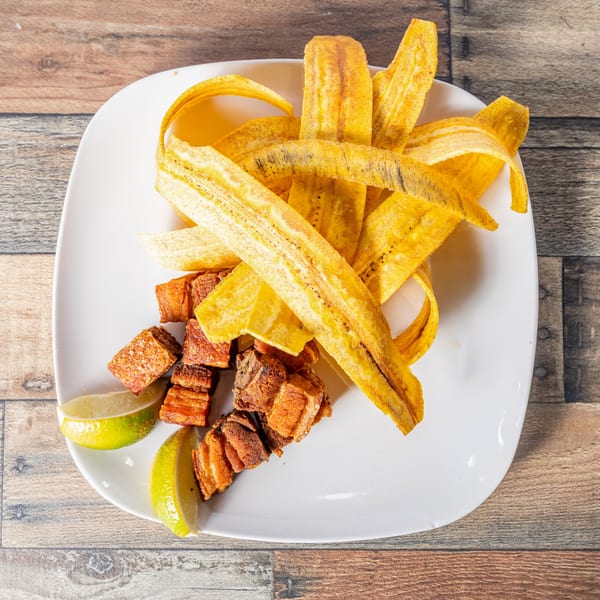 Crispy plantain chips and fried pork belly chunks with lime wedges on a white plate.