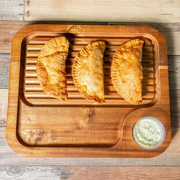 Three golden-brown Cuban empanadas and a small cup of green dipping sauce on a wooden serving board.